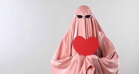 Photo of Cute ghost. Woman in pink sheet with sunglasses and red heart on light grey background, space for text