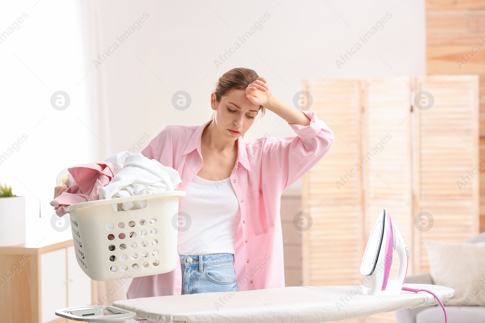 Photo of Young tired woman holding basket of clean laundry at ironing board indoors