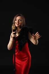 Beautiful young woman in stylish dress with microphone singing on black background