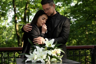 Photo of Sad couple mourning near granite tombstone with white lilies at cemetery. Funeral ceremony