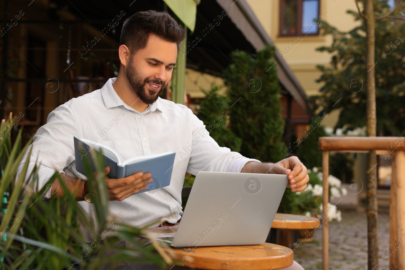 Photo of Handsome young man with book working on laptop at table in outdoor cafe
