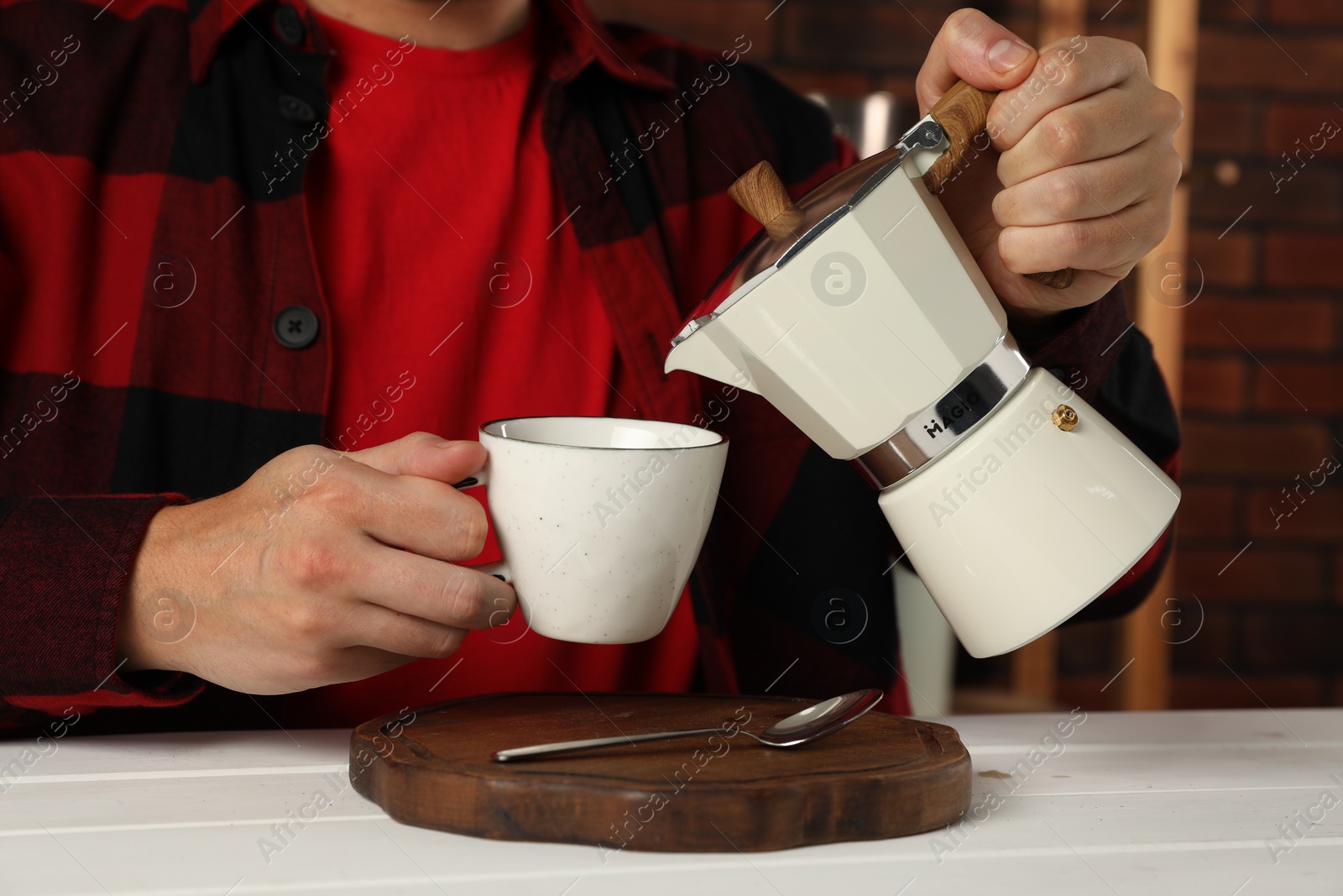 Photo of Man pouring aromatic coffee from moka pot into cup at white wooden table indoors, closeup