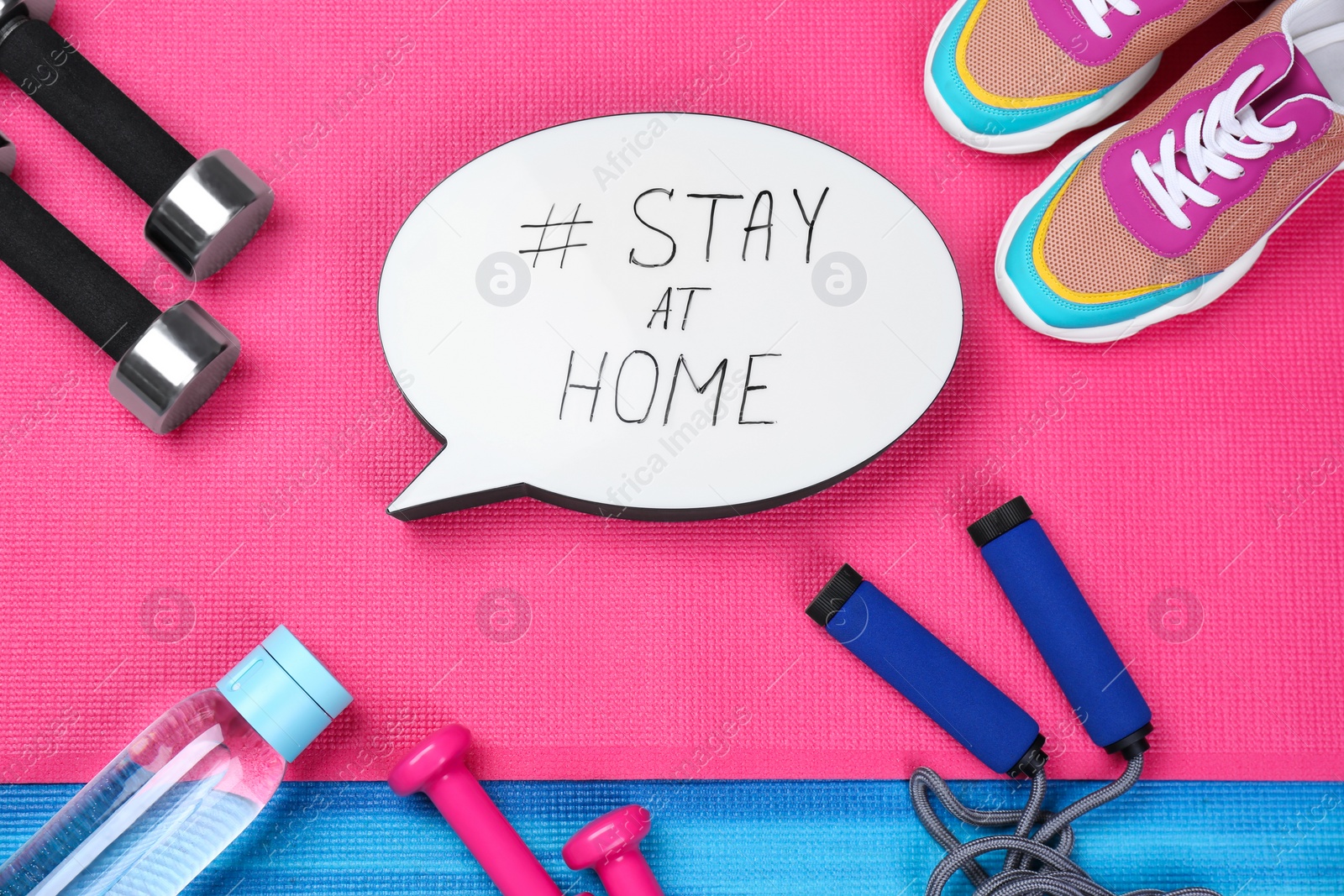 Photo of Sport equipment and speech bubble with hashtag STAY AT HOME on colorful yoga mats, flat lay. Message to promote self-isolation during COVID‑19 pandemic