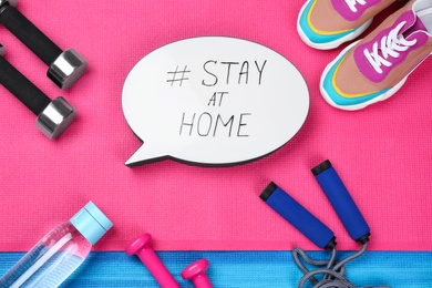 Sport equipment and speech bubble with hashtag STAY AT HOME on colorful yoga mats, flat lay. Message to promote self-isolation during COVID‑19 pandemic