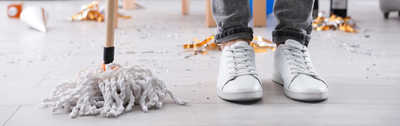 Image of Young man with mop cleaning messy room after party, closeup of legs. Banner design