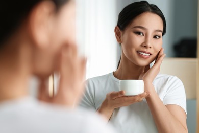 Happy woman applying face cream near mirror at home