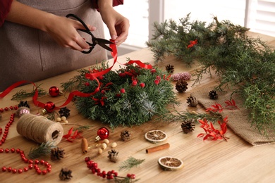 Photo of Florist making beautiful Christmas wreath with berries and red ribbon at wooden table indoors, closeup
