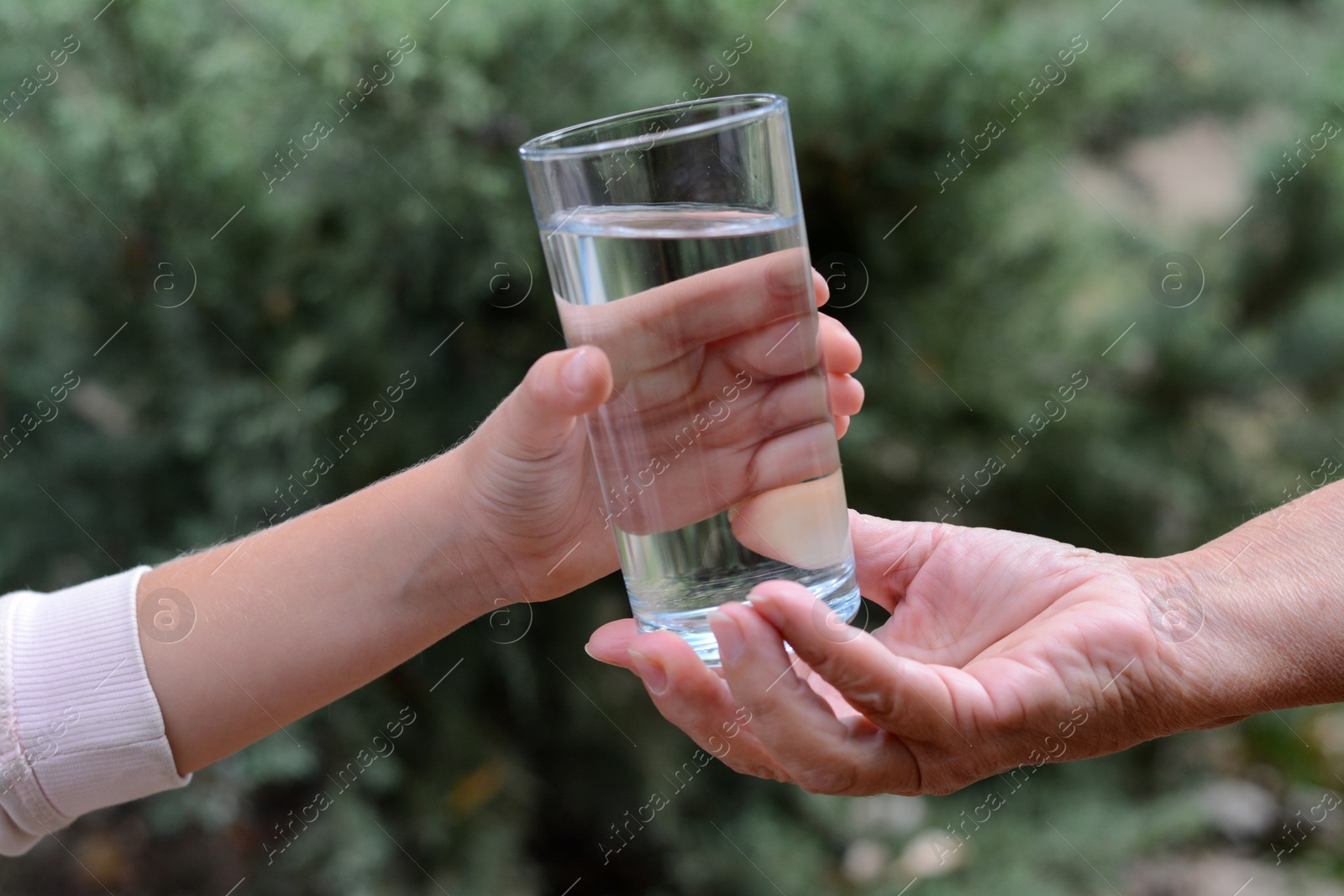 Photo of Child giving glass of water to elderly woman outdoors, closeup