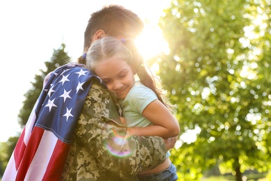 Photo of Father in military uniform with American flag holding his little daughter at green park