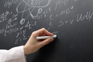 Teacher writing physical formulas with chalk on black chalkboard, closeup