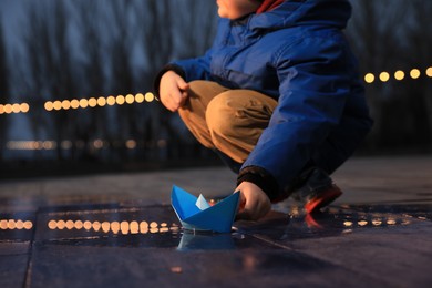 Photo of Little boy playing with paper boat near puddle outdoors, closeup