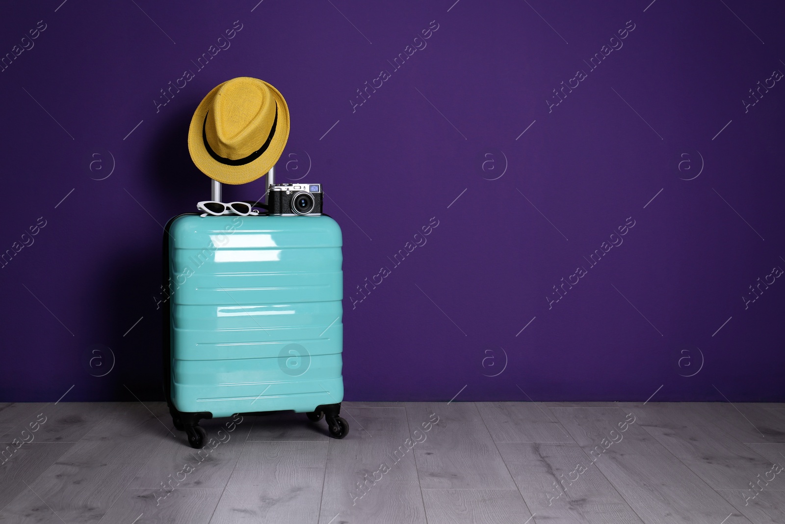 Photo of Travel suitcase with hat, camera and sunglasses on wooden floor near purple wall, space for text. Summer vacation