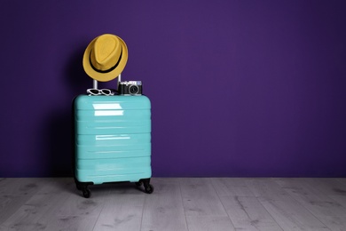 Photo of Travel suitcase with hat, camera and sunglasses on wooden floor near purple wall, space for text. Summer vacation