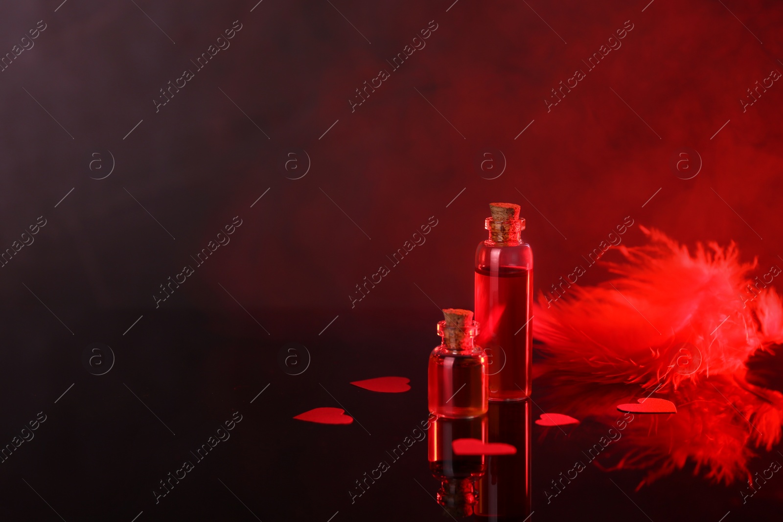 Photo of Bottles of love potion, paper hearts and feather on mirror surface against dark background, space for text. Red tone effect