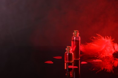 Photo of Bottles of love potion, paper hearts and feather on mirror surface against dark background, space for text. Red tone effect