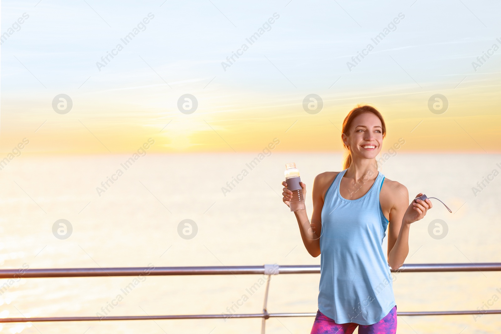 Photo of Young woman holding bottle with water on pier. Morning workout