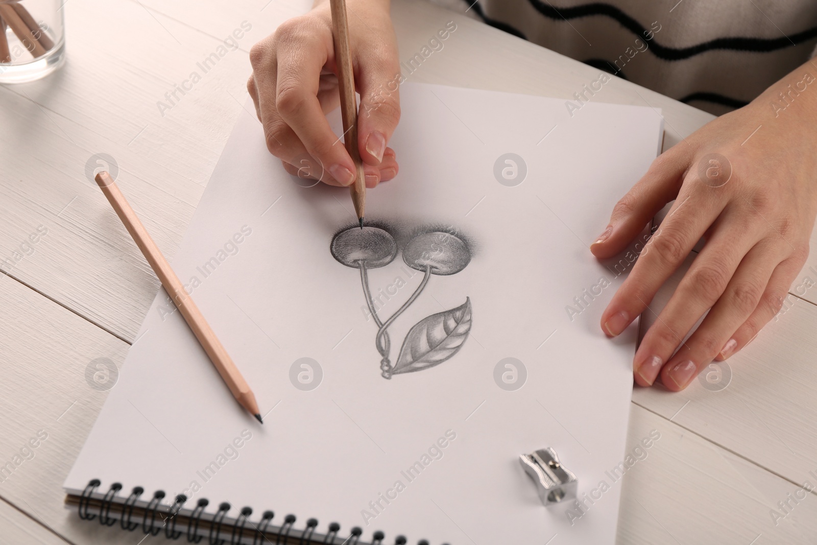 Photo of Woman drawing cherries with graphite pencil in sketchbook at white wooden table, closeup