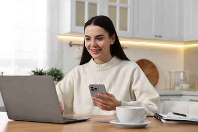 Photo of Happy young woman with gadgets shopping online at wooden table in kitchen