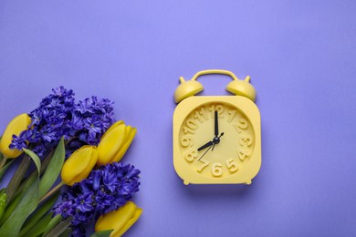 Yellow alarm clock and beautiful flowers on violet background, flat lay. Spring time