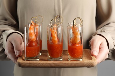 Woman holding tasty canapes with shrimps, tomatoes and sauce in shot glasses on grey background, closeup