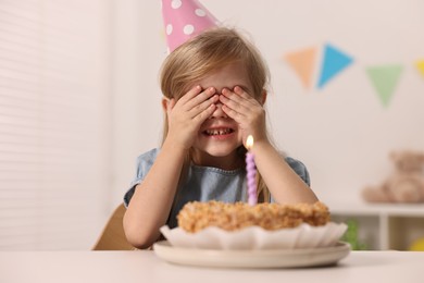 Photo of Cute girl in party hat with birthday cake at table indoors