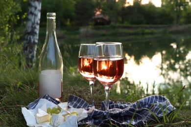 Photo of Delicious rose wine, cheese and grapes on picnic blanket near lake