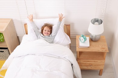 Photo of Cute little boy stretching in cosy bed near alarm clock on bedside table indoors