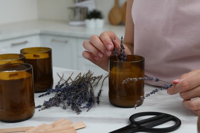 Photo of Woman decorating homemade candle with lavender flowers at table indoors, closeup