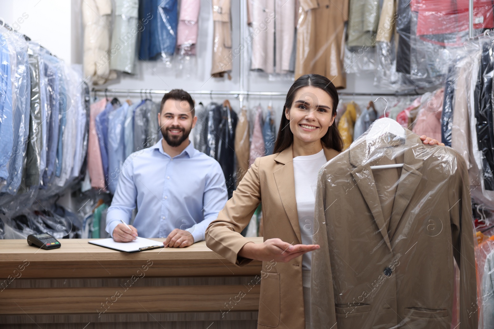 Photo of Dry-cleaning service. Happy woman holding hanger with coat indoors. Worker taking notes at workplace