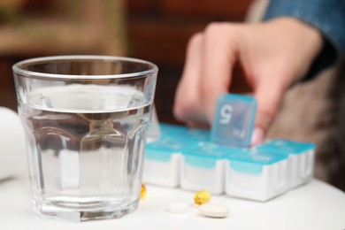 Photo of Woman taking pill from plastic box at white table indoors, focus on glass of water. Space for text