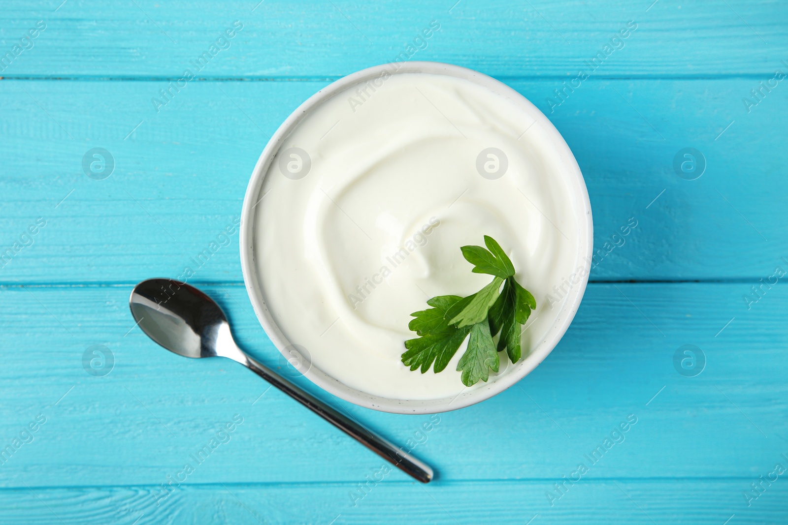 Photo of Bowl of fresh sour cream with parsley and spoon on light blue wooden table, flat lay