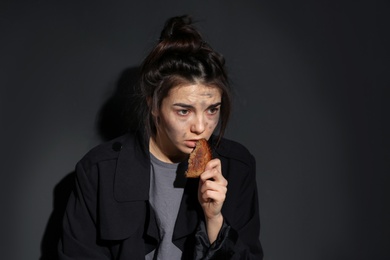 Poor woman with piece of bread on dark background, space for text