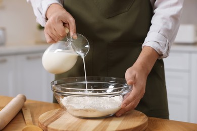 Photo of Making bread. Man pouring milk into bowl with flour at wooden table in kitchen, closeup