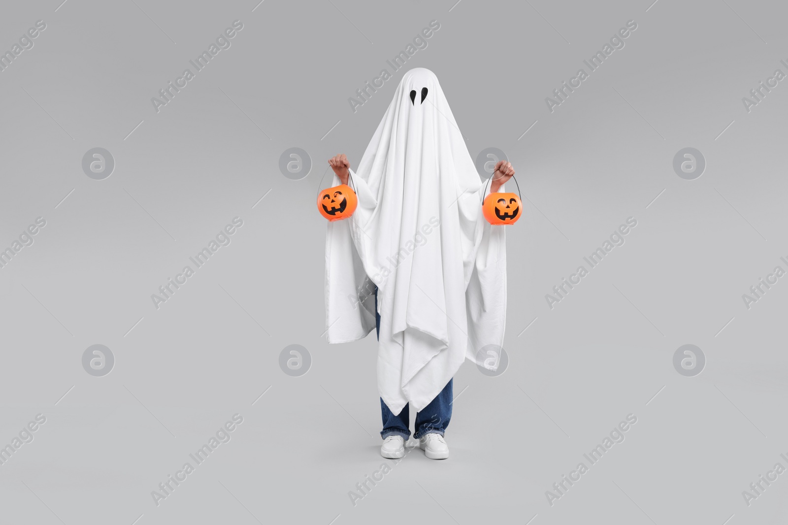 Photo of Woman in white ghost costume holding pumpkin buckets on light grey background. Halloween celebration
