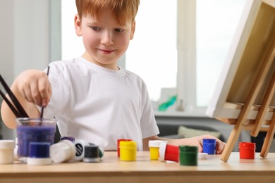 Photo of Little boy painting at table in studio. Using easel to hold canvas