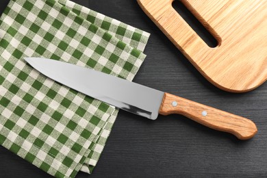 One sharp knife, napkin and board on black wooden table, flat lay