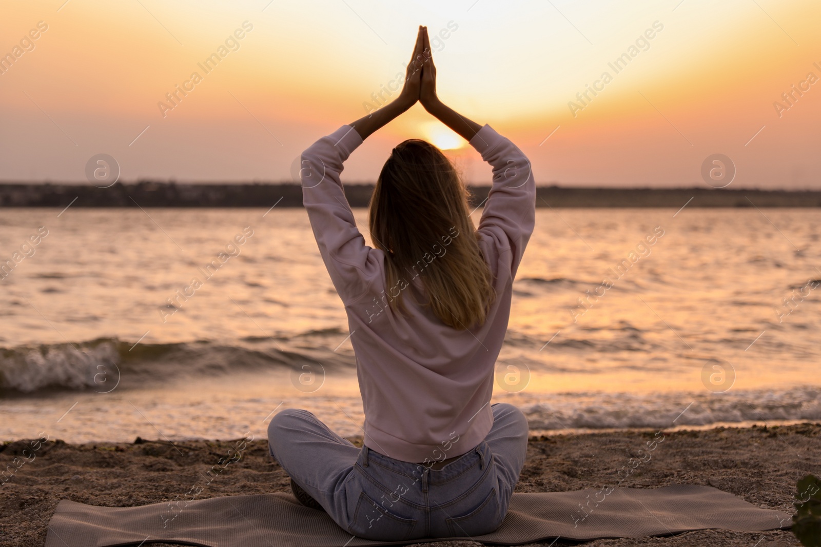 Photo of Young woman meditating on beach near river at sunset, back view