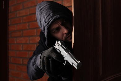 Man with gun spying behind open door indoors. Criminal offence