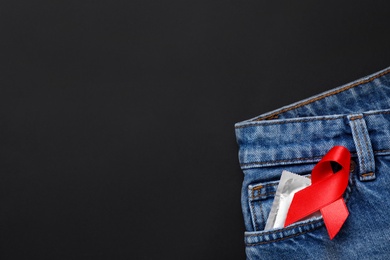 Photo of Top view of jeans with condom and red ribbon in pocket on black background, space for text. AIDS disease awareness