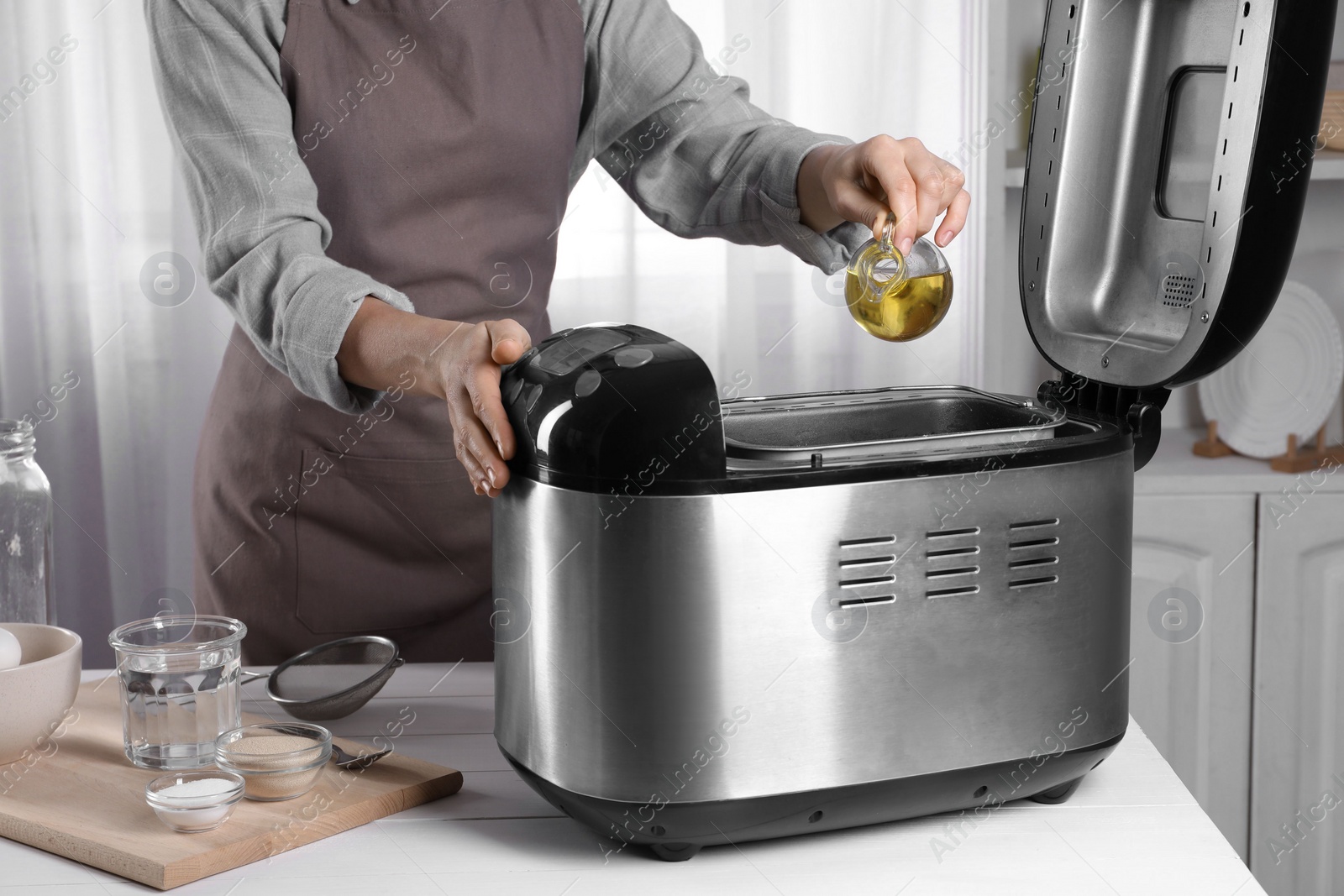 Photo of Woman pouring oil into breadmaker at white wooden table indoors, closeup