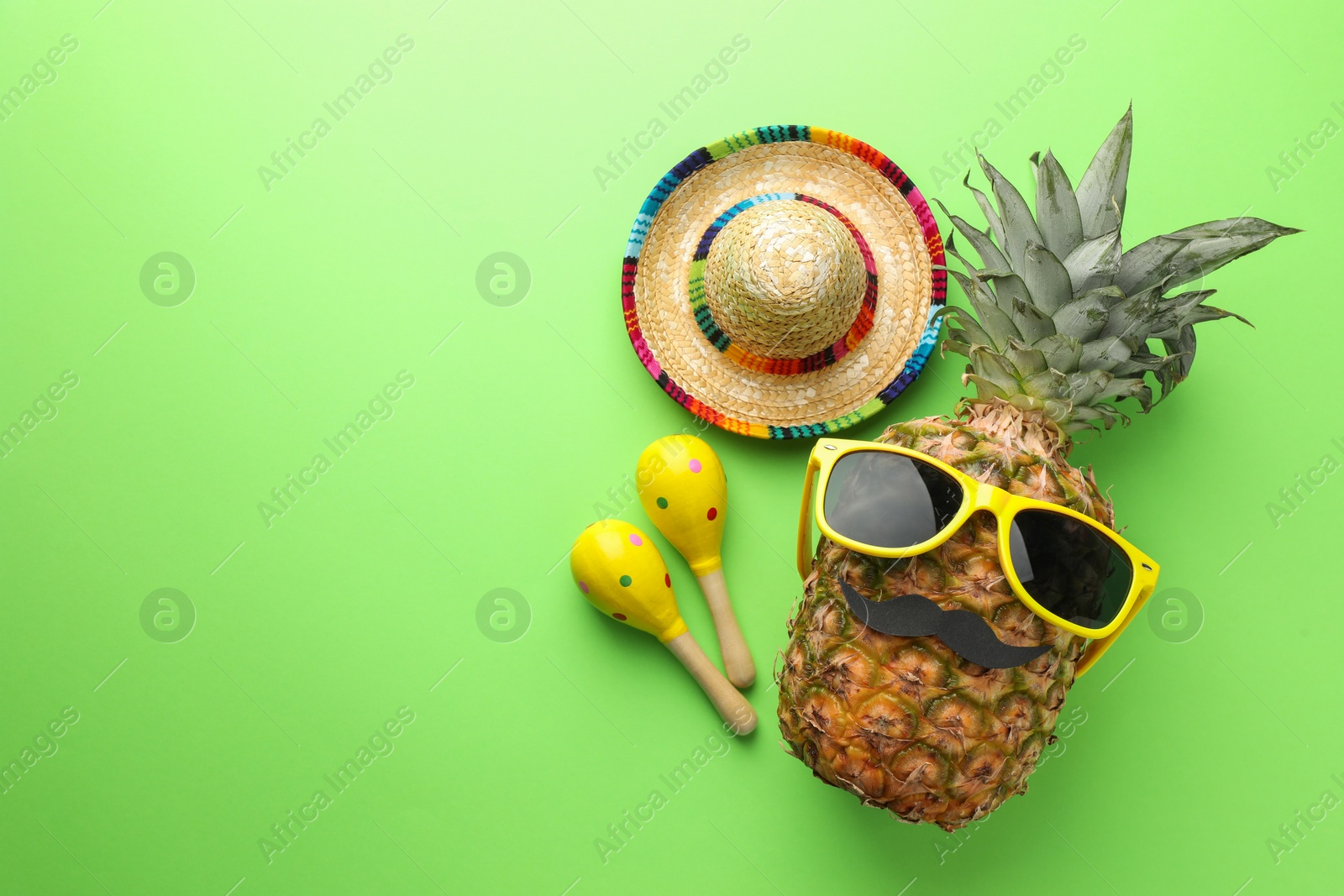 Photo of Mexican sombrero hat, pineapple with sunglasses, fake mustache and maracas on green background, flat lay. Space for text