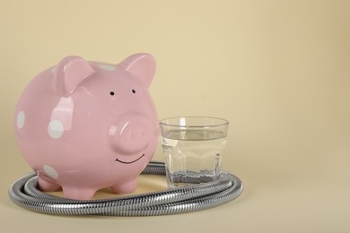 Photo of Water scarcity concept. Piggy bank, shower hose and glass of drink on beige background, space for text