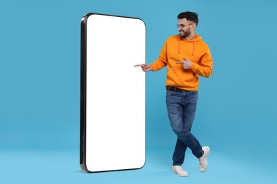 Image of Man pointing at huge mobile phone with empty screen on light blue background. Mockup for design