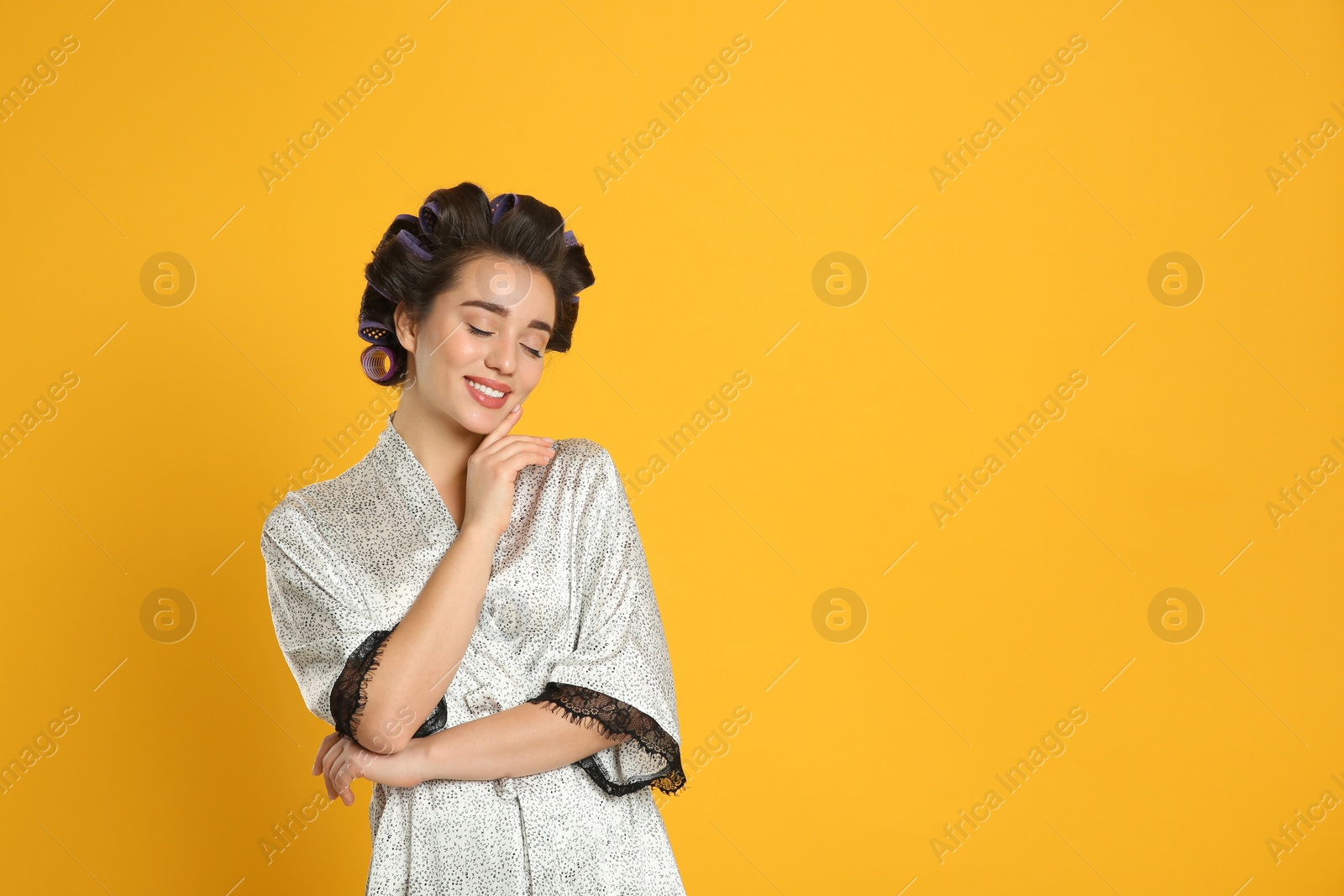 Photo of Happy young woman in silk bathrobe with hair curlers on orange background, space for text