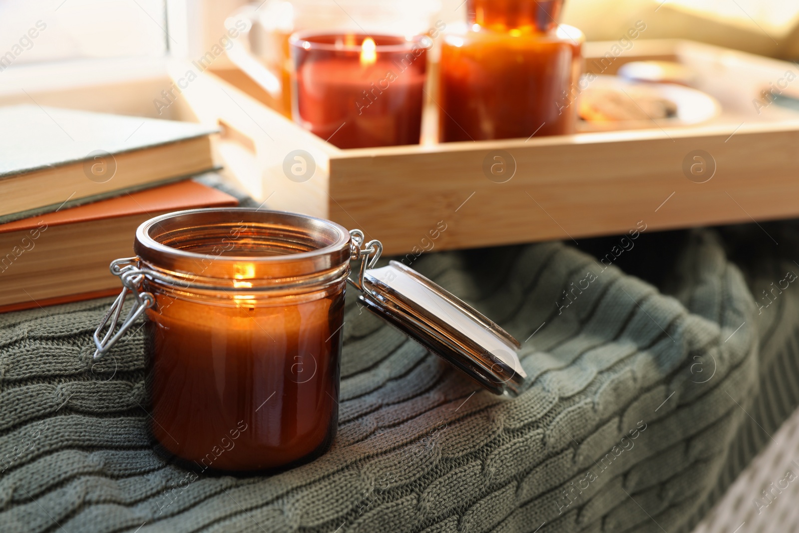 Photo of Burning candle in jar on knitted plaid near window indoors