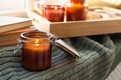 Burning candle in jar on knitted plaid near window indoors