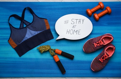 Photo of Sport equipment and speech bubble with hashtag STAY AT HOME on blue yoga mat, flat lay. Message to promote self-isolation during COVID‑19 pandemic