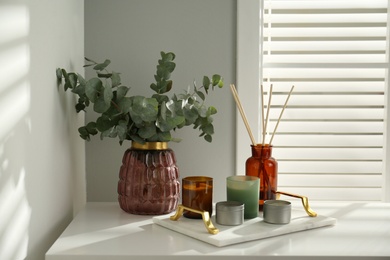 Photo of Eucalyptus branches, aromatic reed air freshener and candles on white table near window indoors. Interior element