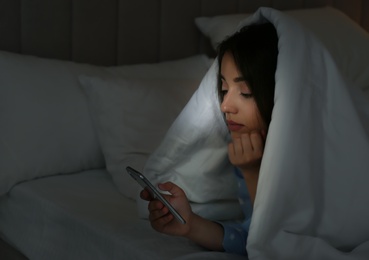 Photo of Young woman using mobile phone under blanket in dark bedroom