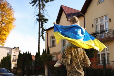 Photo of Soldier in military uniform carrying his daughter on shoulders and Ukrainian flag outdoors, space for text. Family reunion